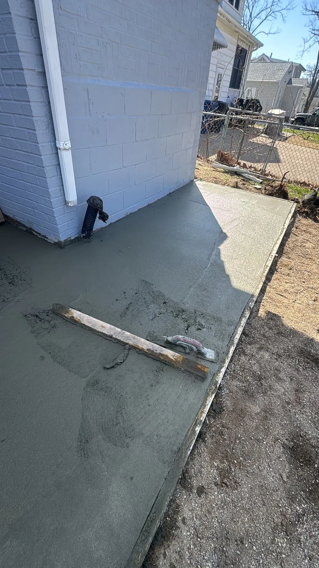 Fresh concrete walkway against a white brick wall, with the screed board and float still on it.